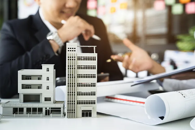Architectural models of two apartment buildings on a desk with rolled plans, a man in a suit blurred in the background
