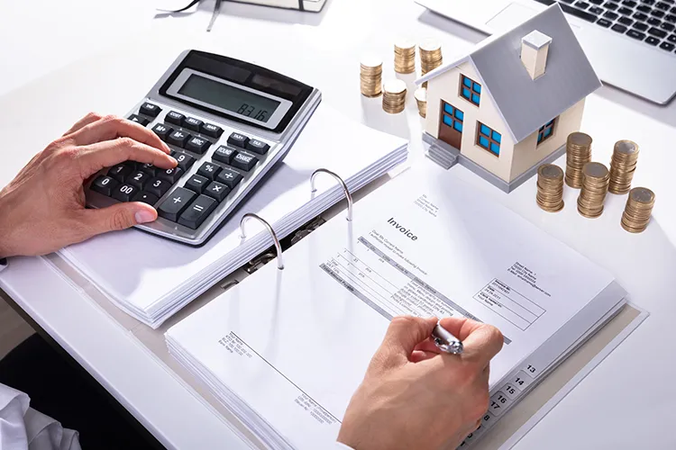 Hand on a calculator beside an invoice in a ring binder, a model house and stacks of coins on a desk