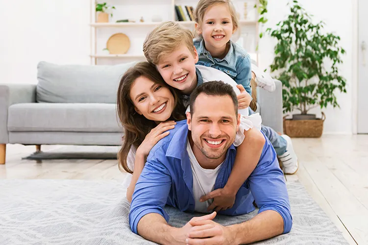 Parents and two young children lying in a smiling pile on a living room rug in front of a grey sofa