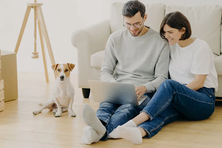 Couple sitting on the floor with a laptop in front of a white sofa, a small white and tan dog and moving boxes beside them