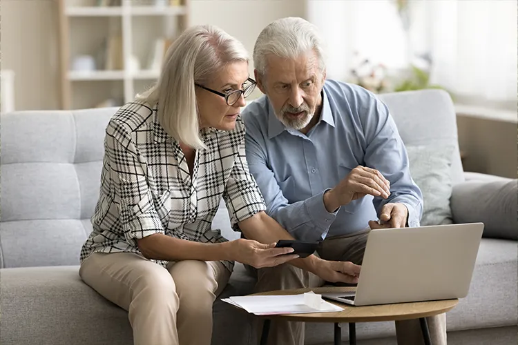 Older couple on a sofa studying a laptop and calculator, with papers on the coffee table in front of them