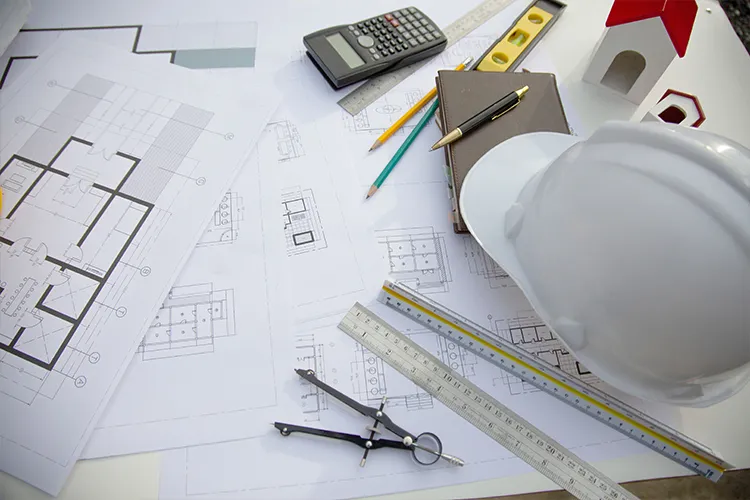 Architectural floor plans spread on a desk with a white hard hat, calculator, rulers, drawing compass and a model house