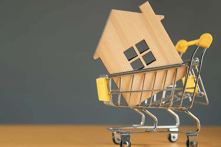 Wooden cut-out house sitting inside a miniature shopping trolley on a wooden table